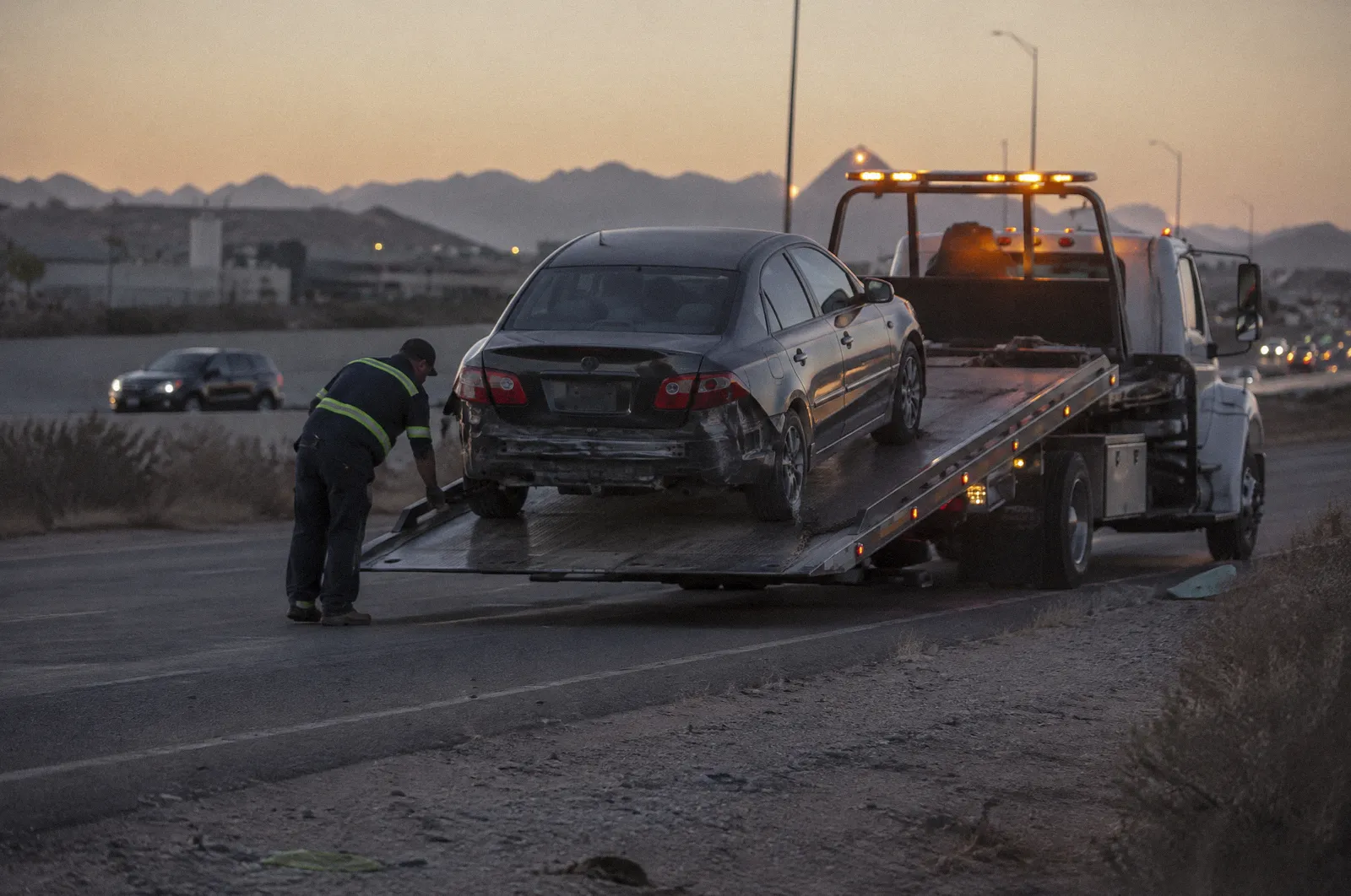 A flatbed tow truck loading a damaged car at dusk, amber lights flashing, freeway frontage road