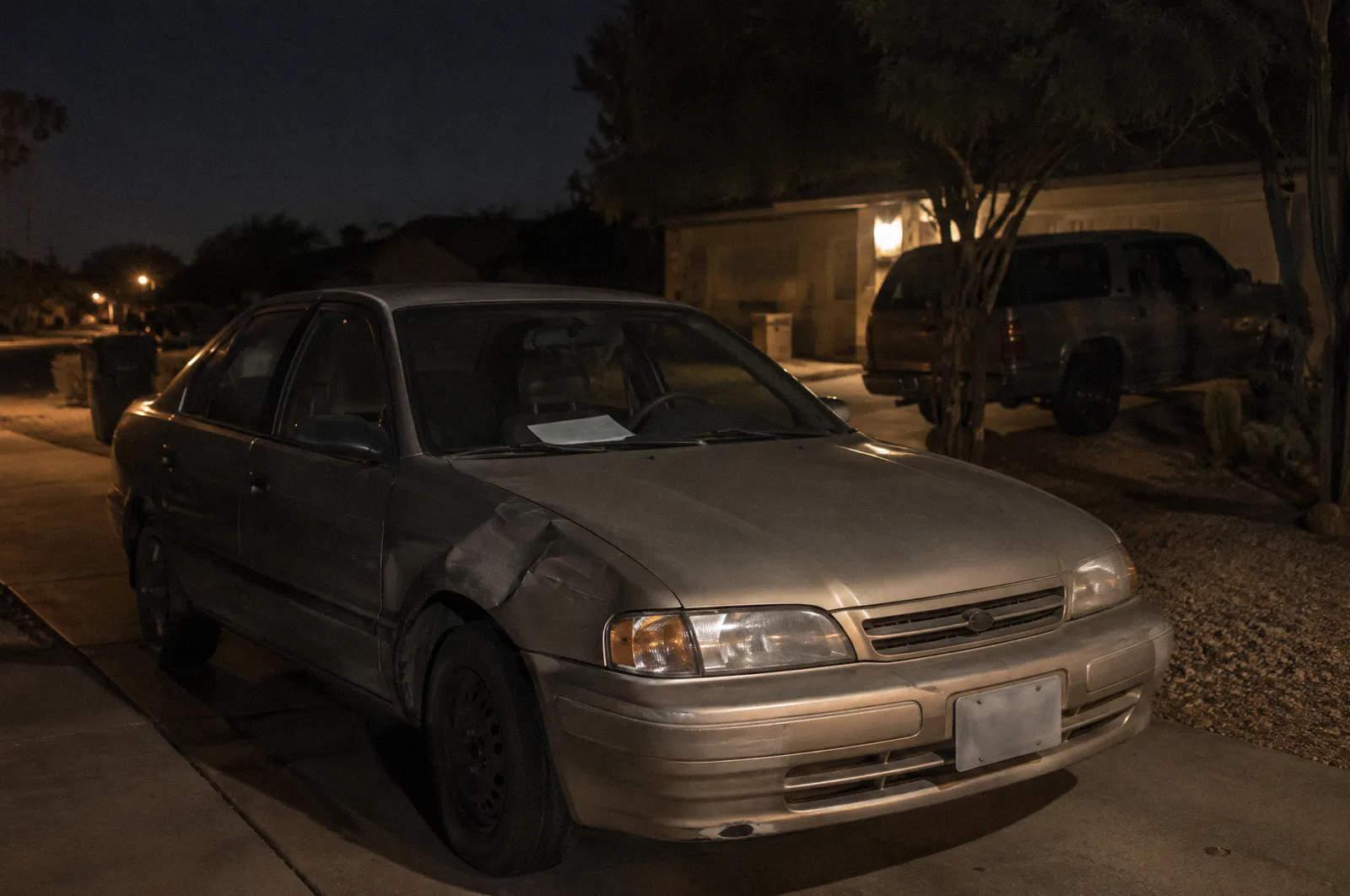 A dented older car parked in a driveway with a blank paper on the dash, harsh noon light