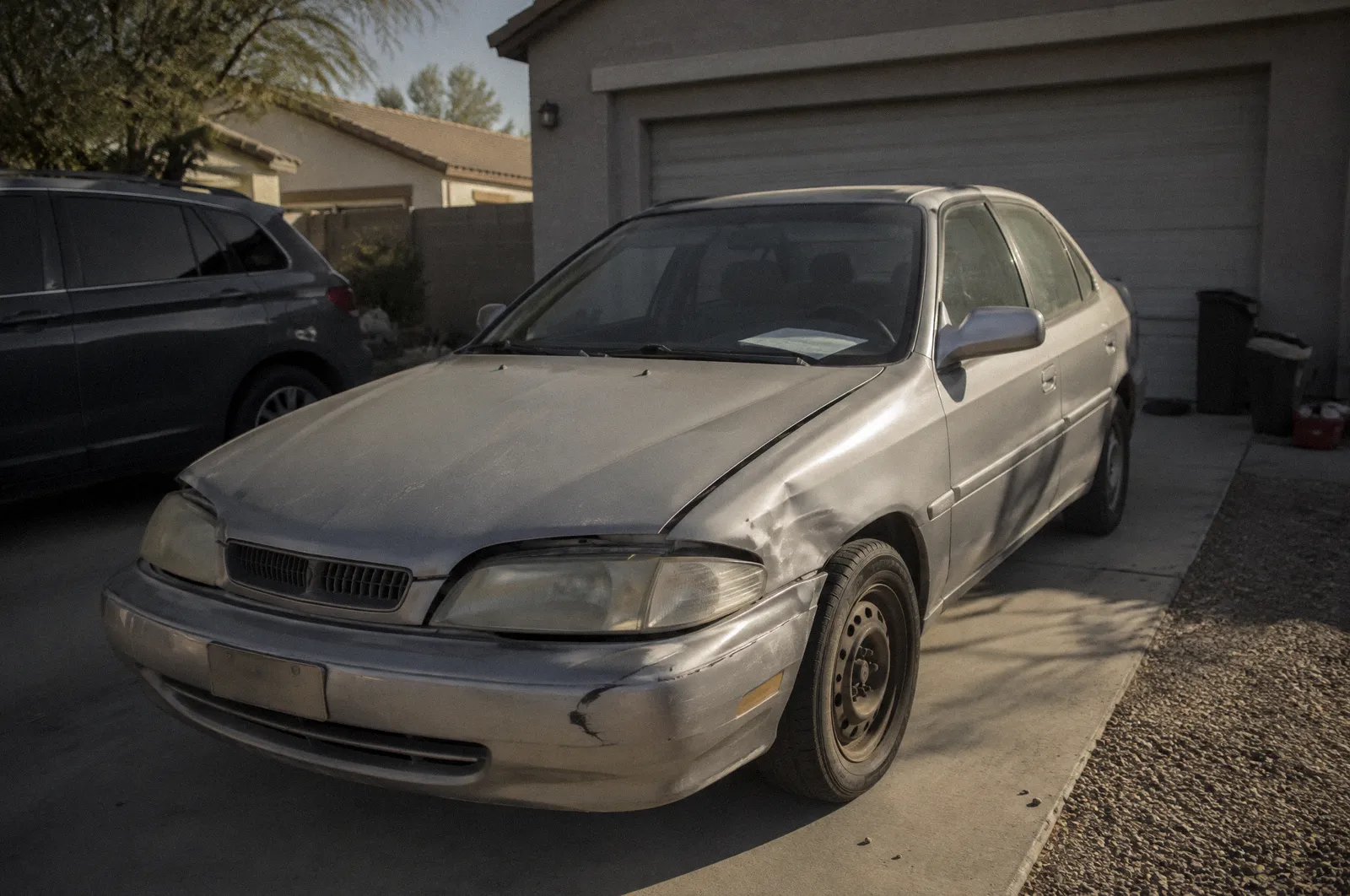 A dented older car parked in a driveway with a blank paper on the dash, harsh noon light
