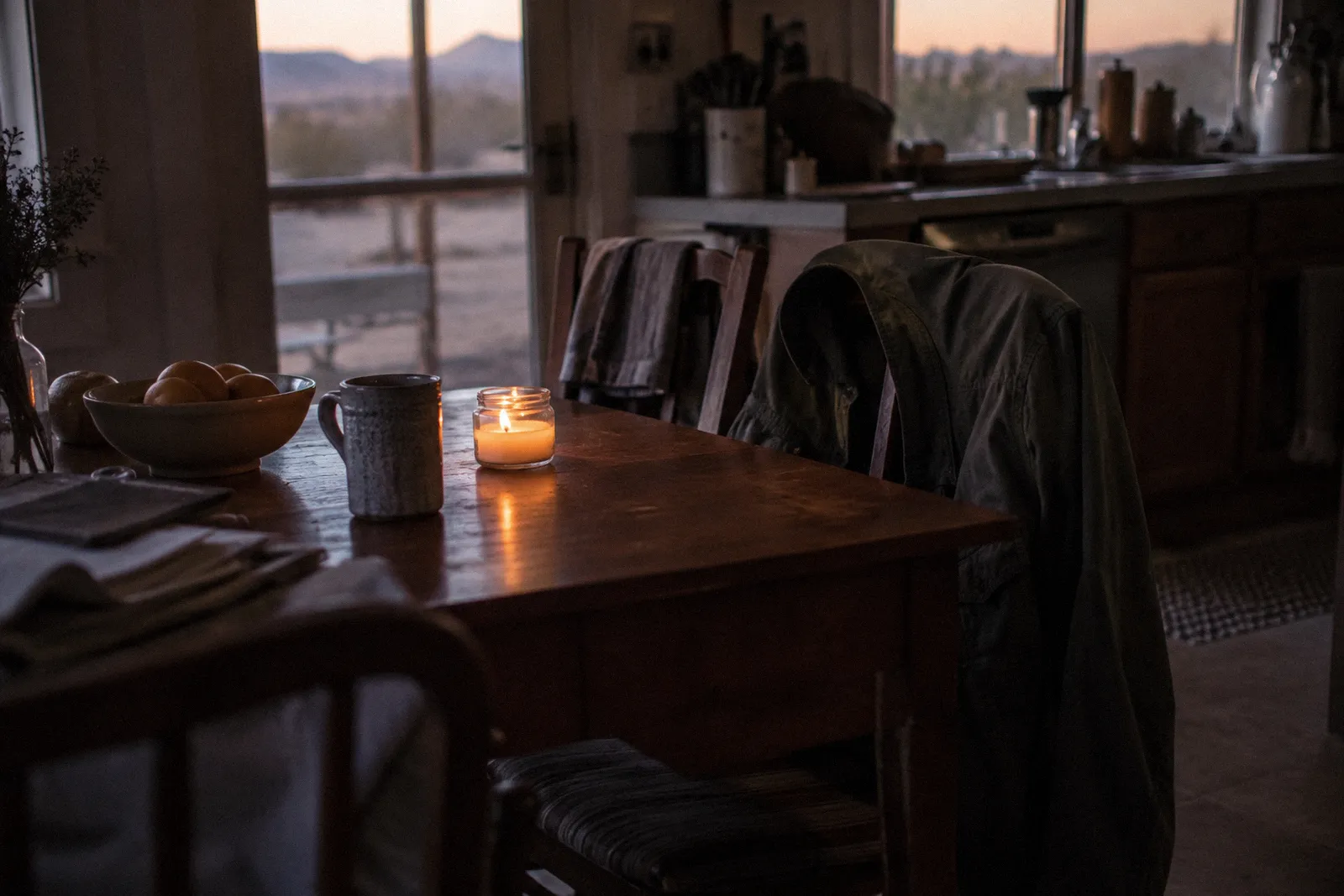 A single lit candle on a kitchen table beside a folded jacket on an empty chair, soft evening light