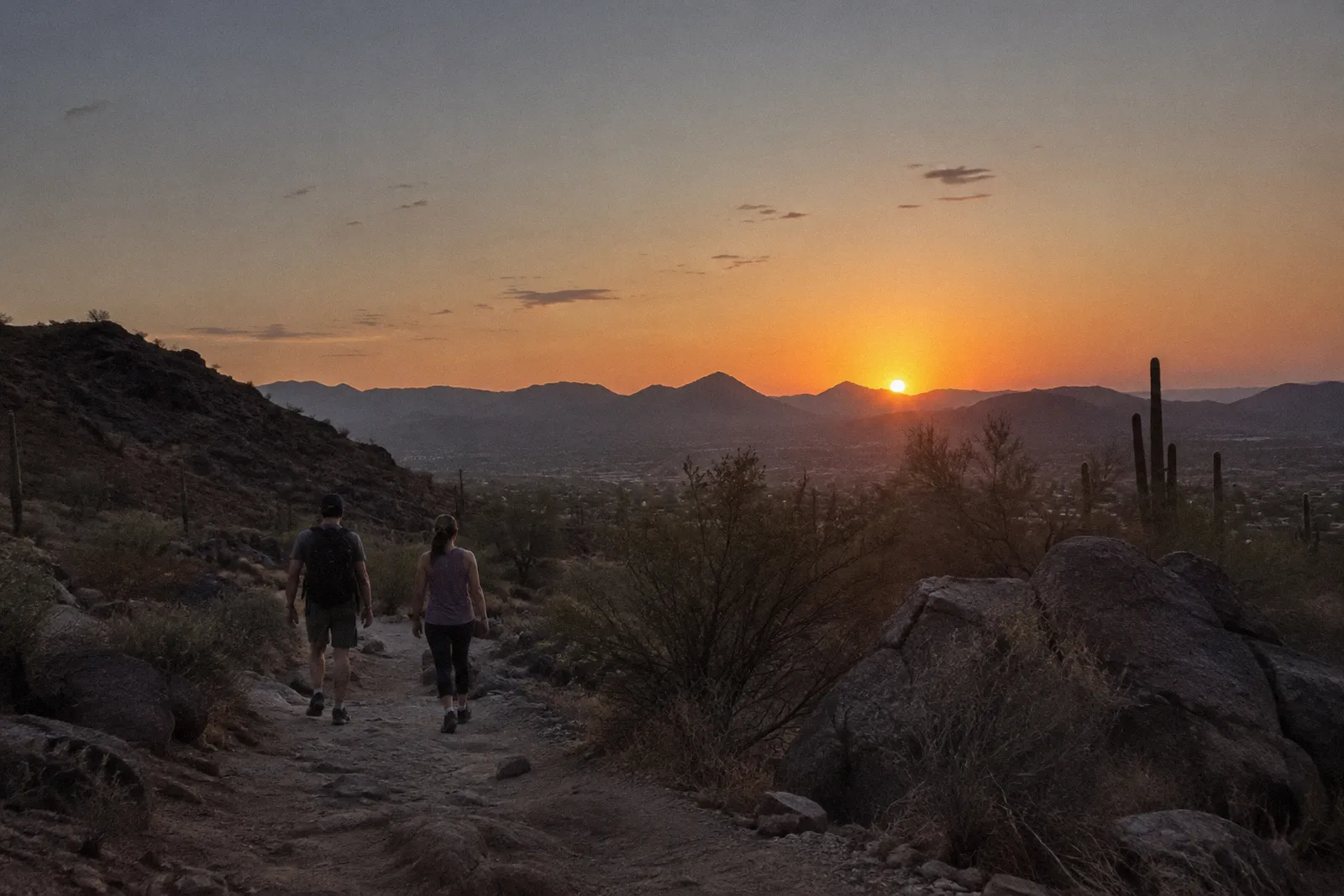 Two adults seen from behind walking slowly on a desert trail at sunset, small in the frame